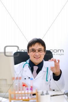 Smiling doctor sitting at office table and holding blank business card
