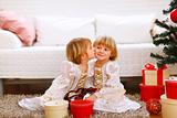 Twin girl kissing her sister near Christmas tree with gifts
