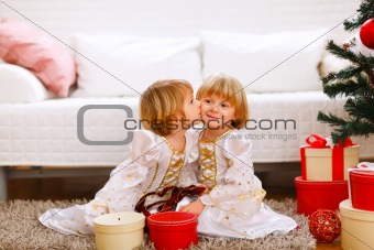 Twin girl kissing her sister near Christmas tree with gifts
