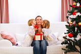 Two twins daughters presenting gift to mother and kissing her near Christmas tree
