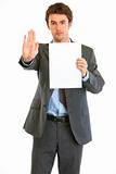 Serious modern businessman holding blank paper and showing stop gesture
