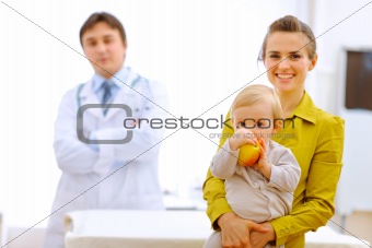 Portrait of mother with baby holding apple and doctor in background
