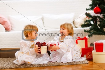 Two lovely twins girl exchanging presents near under Christmas tree