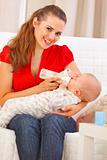 Smiling young mother sitting on couch and feeding baby