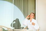 Laughing young woman in bathrobe with cup of coffee speaking mobile phone on terrace