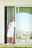 Beautiful woman in bathrobe standing on terrace near big windows and having cup of coffee