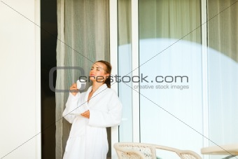 Lovely woman in bathrobe standing near big room windows and having cup of coffee
