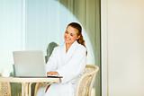 Smiling young woman in bathrobe sitting at table on terrace and using laptop