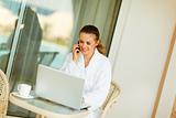 Happy woman in bathrobe sitting at table on terrace speaking mobile and looking in laptop