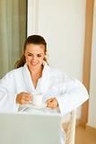 Happy woman in bathrobe sitting on terrace having coffee and exploring something in notebook