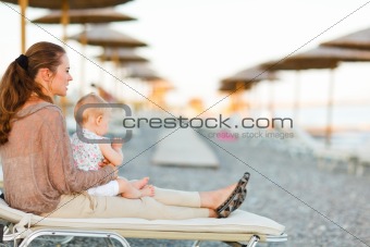 Young mother sitting with baby on sunbed on the beach at evening and looking into the distance