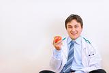 Smiling medical doctor sitting on floor and holding apple in hand