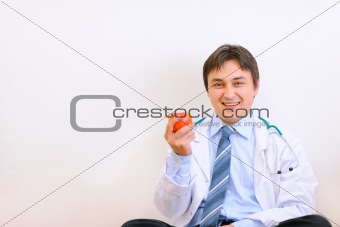 Smiling medical doctor sitting on floor and holding apple in hand
