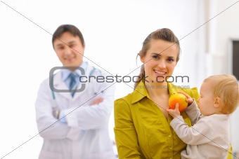 Portrait of mother with baby holding apple and doctor in background