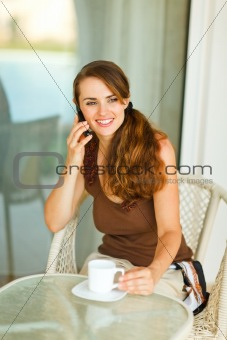 Happy young woman speaking cell phone and having cup of coffee at terrace