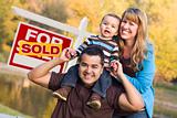 Happy Mixed Race Couple with Baby in Front of Sold Real Estate Sign.