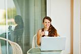 Laughing young woman enjoying cup of coffee and looking in laptop