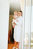 Smiling mother in bathrobe with baby looking out from window