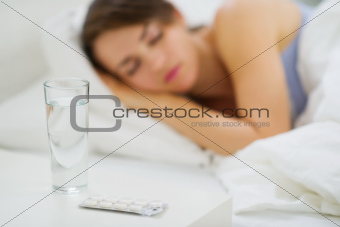 Closeup on pills and glass of water on table and sleeping woman in background