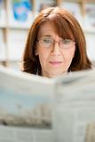 Elderly woman with glasses reading newspaper in library