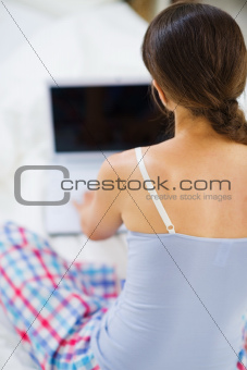 Woman sitting on bed and working on laptop. Rear view