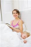 Smiling mother in bed reading book while baby sleeping