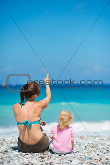 Mother sitting with baby on beach and pointing on copy space. Rear view