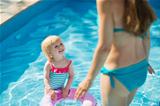 Baby standing in pool with inflatable ring and looking on mother