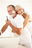 Senior Man Shaving In Bathroom Mirror With Wife Watching