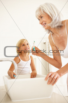 Grandmother Brushing Teeth In Bathroom With Granddaughter Watching