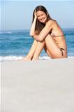 Young Woman Sitting On Sandy Beach On Holiday Wearing Bikini