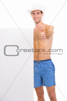 Happy young man in shorts and hat showing blank billboard and thumbs up
