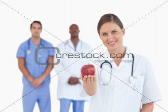 Smiling doctor offering apple with colleagues behind her