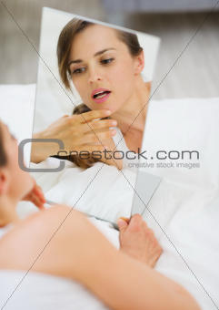 Young woman laying on bed and checking face in mirror