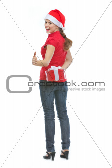 Happy young woman hiding Christmas present box behind back and showing thumbs up