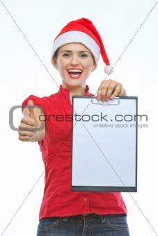 Happy young woman in Christmas hat showing blank clipboard and thumbs up