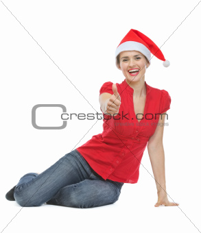 Happy woman in Christmas hat sitting on floor and showing thumbs up