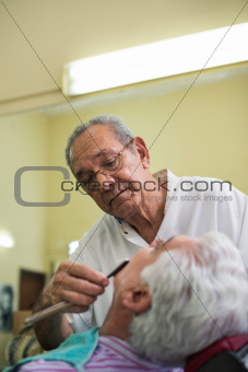 Elderly barber with razor shaving client in barber shop