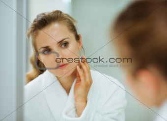 Young woman in bathrobe checking her face in mirror in bathroom