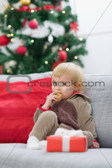 Baby in suit of Santa's little helper with Christmas gift eating cookie