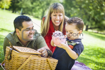 Happy Mixed Race Couple Give Their Son a Piggy Bank at a Picnic in the Park.