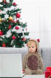 Baby in Christmas deer costume near Christmas tree looking in laptop