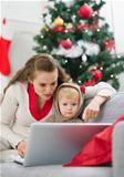 Mother showing baby something in laptop near Christmas tree