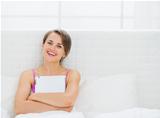 Happy young woman sitting on bed and embracing tablet PC