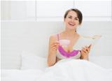 Happy young woman with book and cup in bed