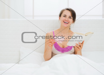 Happy young woman with book and cup in bed