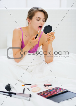Young woman sitting in bed and applying makeup