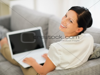 Smiling young woman laying on couch and working on laptop