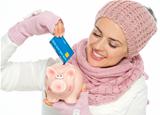 Smiling woman in knit scarf, hat and mittens putting credit card in piggy bank