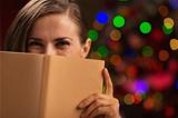 Happy woman hiding behind book in front of Christmas lights